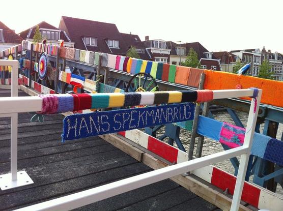 Brug over het water, waarlangs huizen staan. De brugleuningen zijn met breiwerkjes versierd, op het voorste hekje is een gebreid bordje met de tekst 'Hans Spekmanbrug' zichtbaar
