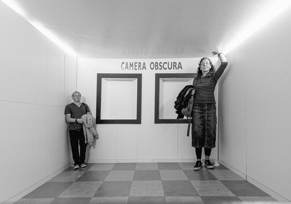 Schwarzweissfotografie eines Raumes mit zwei Personen in je einer Ecke; die Grössenverhältnisse sind verzerrt. 
