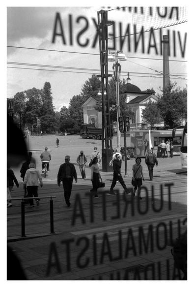 A view of the Tampere Central Market Square, shot through a window glass of a hamburger place. People crossing the tram rails on green light, the Old Church is visible beyond the tram electric poles.