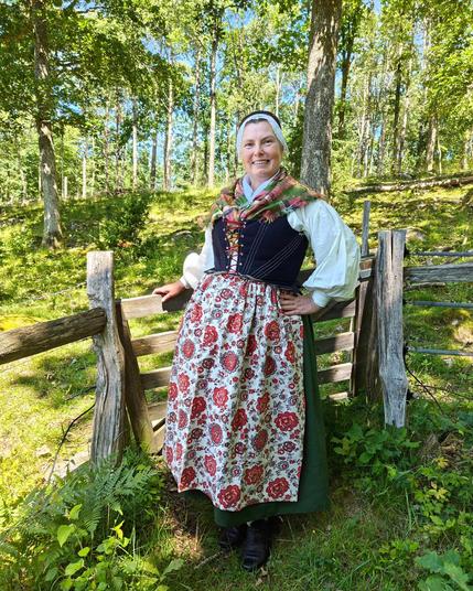 A white middle aged woman is leaning against a wooden gate in a wooded field. She is wearing a white shirt, a dark blue bodice, a green skirt, which is hardly visible beneath a large flowers apron. She has a checked silk scarf over her shoulders, and a black cap with white lace in front.