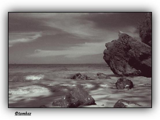 Black-and-white seaside landscape featuring a rocky shoreline. In the foreground, several large, irregularly shaped rocks protrude from the water, their rough, jagged texture visible. The sea is calm, with gentle waves softly blending into the shore. Sunlight reflections create shimmering streaks on the water’s surface, enhancing the contrast between the light and dark areas of the image. In the background, the open expanse of the sea meets the horizon, where a faint coastline adds depth to the composition. The sky is covered in soft, elongated clouds, forming a dynamic yet tranquil backdrop for the entire scene.