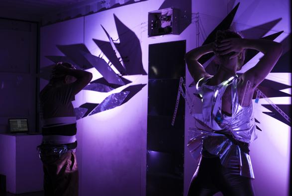 Two people under purple lighting, facing away from the camera with their hands on the back of their heads. The person closest to the camera has mechanical-looking 'wings' on their back.