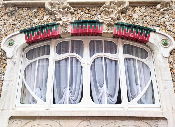 Ein Jugendstil-Fenster mit einem bogenartigen Fensterrahmen, durchsetzt mit roten und grünen Ziegeldekorationen, die Fenster mit durchweg runden Sprossen, dahinter weiße Vorhänge. 