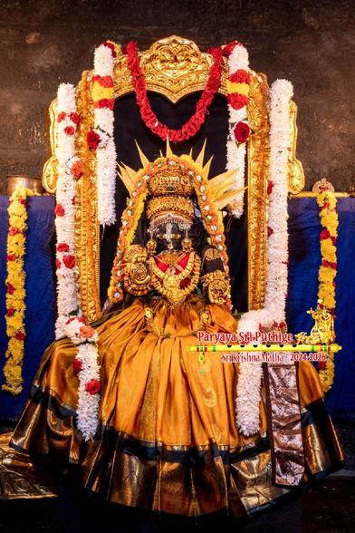 Idol of Hindu diety Krishna dressed in light brown (burned aubergine/eggplant) saree with feminine appearance, seated with the saree pleated and spread wide, wearing gold crown, and several gold ornaments. Yellow petals of kaetaki arranged behind the crown to form an aura. A string of orange roses and a string of yellow mums both go up from Krishna's left waist, over the crown, and then down to the right waist.

Gold frame/arch behind Krishna. Two thick strings of white flowers, one on either arm of the frame, drop from the top of the frame, flows down and Krishna's thighs, and drops down in his front.

A short string of red roses in the middle top of the frame.

Blue cloth background.