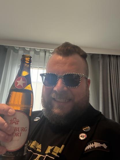 A smiling individual with a beard is holding a bottle of beer while wearing black and white checkered sunglasses. They are dressed in a dark jacket with several pins and have a casual black t-shirt. The background features light-colored curtains.