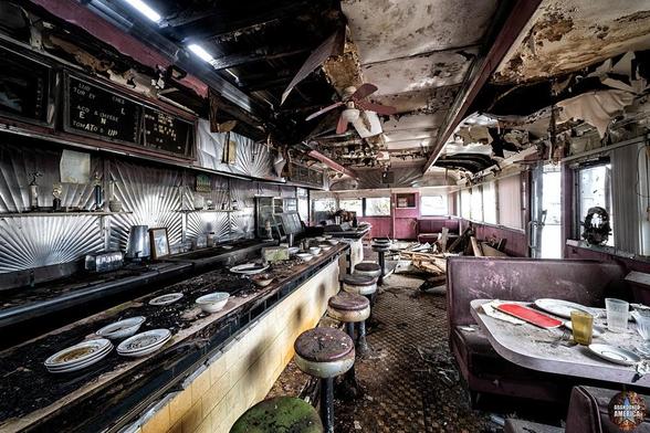 A diner counter covered with debris and dishes is on the left, and a table still set with plates is on the right. The roof is badly damaged and light is peeking through holes in it
