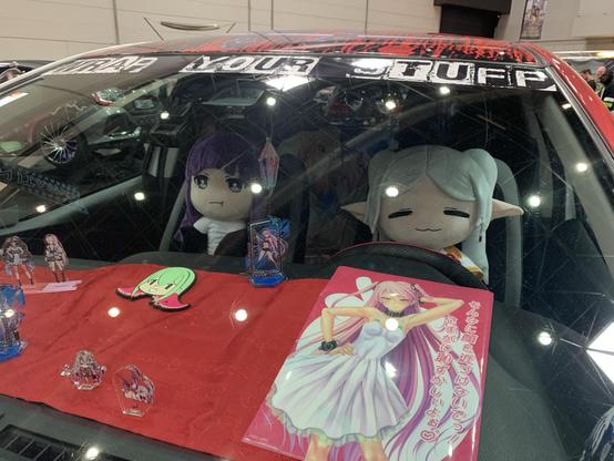 In the photo, Frieren (driver) and Fern are sitting as plush dolls on the front seats of a sports car customized in an anime style.