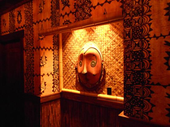 An alcove in a Tiki bar, batik wallpaper, woven rattan backdrop, bamboo framing. There's a dramatically-lit oval mask in the middle. Quite probably not a real Polynesian artifact, but made by Oceanic Arts. 
