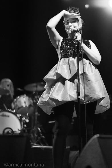 A performer sings into a microphone on stage, wearing a shiny black and silver outfit with a voluminous skirt. The image captures a moment of expression, with the artist gesturing playfully. In the background, a drummer is partially visible. It is a black and white picture.