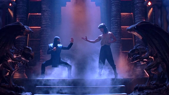 Cena do filme Mortal Kombat (1995). Sub-Zero (François Petit) e Liu Kang (Robin Shou) estão prestes a iniciar um combate intenso. Sub-Zero, com sua clássica vestimenta azul e preta, encara Liu Kang, que veste roupas de luta tradicionais. O ambiente ao redor é sombrio e misterioso, com iluminação dramática que destaca os personagens. A tensão da batalha iminente é evidente na postura dos lutadores.