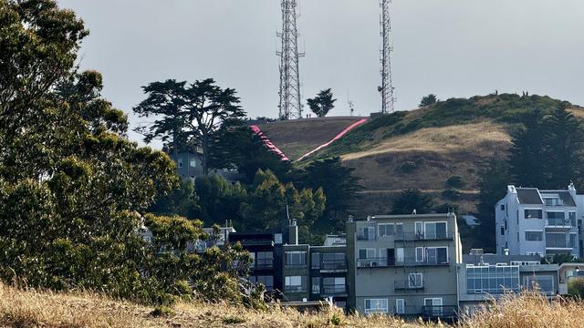 Distant hill in terrible light supporting the pink outline of a giant triangle. Houses and brown grass in the foreground. 