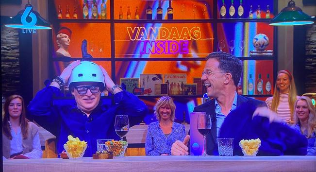 Mark Rutte bij VI, schaterlachend om René van der Gijp die een helm met daarop een dildo op zijn hoofd zet.
