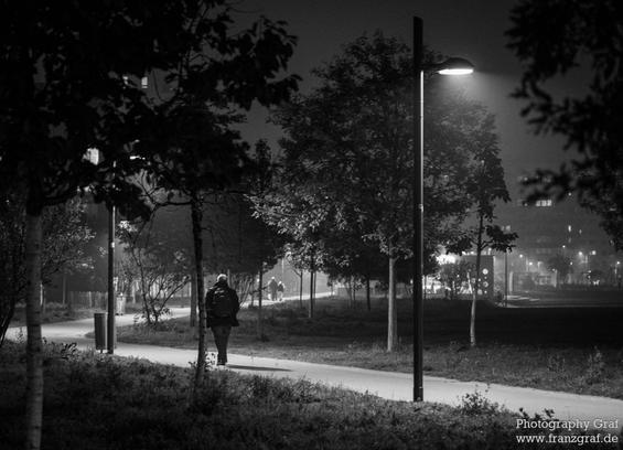 A black and white image of a person walking alone on a sidewalk at night. The person, wearing dark clothing, is captured in mid-stride, highlighted against the dimly lit background. Street lights cast a soft glow, illuminating the scene and creating a mysterious atmosphere. In the background, there are trees and a faint fog adding to the eerie ambiance. The sidewalk is lined with grass, and in the distance, a palm tree is silhouetted against the night sky. The overall mood is peaceful yet sligh…