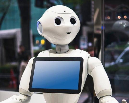 A photo of a Pepper humanoid robot (a white, stylised human-like robot with large eyes and a tablet on its torso). 
