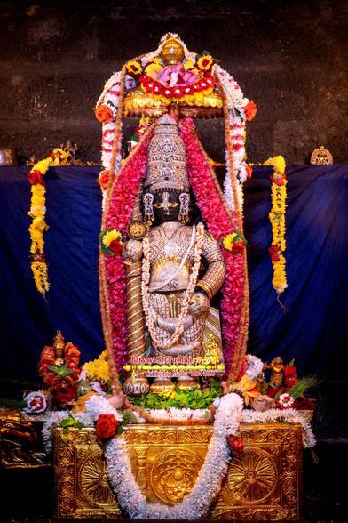 Idol of Hindu deity in diamond cladding and a matching crown, a similarly clad staff in right hand, wearing gold shoes and a long garland of white flowers (with orange stem). A thick dense string of red flowers around Krishna going all the way over the crown.

Krishna standing on an ornate gold box. A thick string of white flowers shaped like a U on the box. A big red rose in both arms of the U. Some green holy basil leaves on the box at Krishna's feet.

A small gold umbrella over Krishna. A thick string of white flowers (with stripes of red flowers) over the umbrella and hanging down on either side of Krishna.

Blue cloth background. A string of yellow flowers (with some spots of red flowers) hanging down from both edges. 