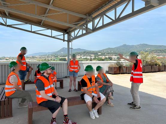 Diverses persones assegudes en bancs amb armilles i cascs prestant atenció al guia explicant.