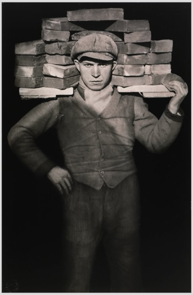 Zwartwitfoto van man die plank met bakstenen draagt. De foto is gemaakt door August Sander in 1928.