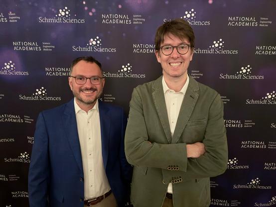 Gabriel Farías and Miguel Dobrich standing side by side, smiling, in front of a National Academies and Schmidt Sciences poster. Both are wearing casual-professional outfits.