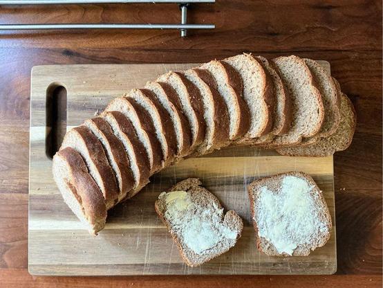 Flat lay view of a sliced loaf of sandwich bread on a wooden cutting board, on top of a dark wooden counter. There are also 2 slices of buttered bread on the board.