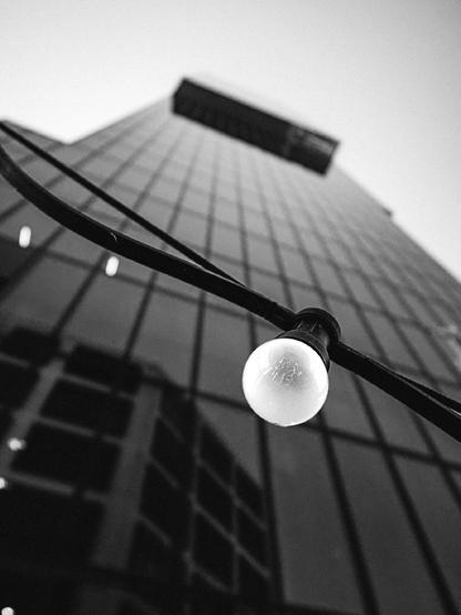 Black and white upward view of a tall glass skyscraper, with a single exposed light bulb from an outdoor fairy light strung diagonally in the foreground. The bulb hangs in sharp focus against the blurred grid of the building’s reflective windows.

