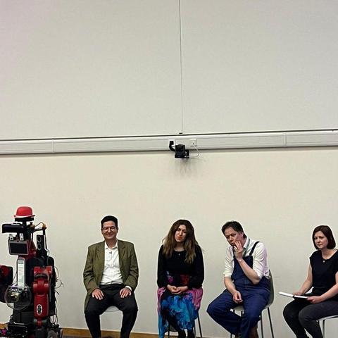 A panel of four people on stage with a red humanoid robot. They’re looking with interest as they listen to a question from the audience.