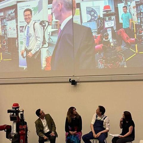 A panel of four people on stage with a red humanoid robot. They’re looking up at the projector screen behind them which shows a man wearing a teleoperation backpack next to the same red robot.