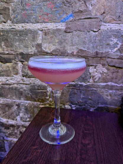 A cocktail in a coup on a table: the cocktail is topped with egg white foam and it's dark red at the top, fading to pale pink at the bottom.