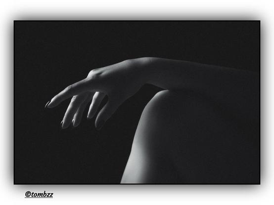 The photo captures an elegant, black-and-white composition focusing on a delicate detail of a woman's silhouette. At the center of the image, a slender leg provides support for a gracefully positioned hand. The woman's fingers form a sensual gesture, subtly emphasizing the softness and fluidity of movement. The contrast of the dark background enhances the mystery of the scene, bringing out the subtle interplay of light and shadow, which gives the composition a deep, elegant character. The play of light and shadow adds depth to the image, creating a magical, refined atmosphere.