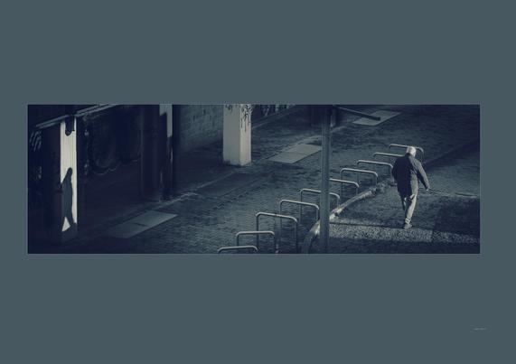 A man walks slowly away from warm sunlight, his long shadow cast diagonally onto a distant pillar. The subtle dislocation—him in one corner, his shadow in another—creates a surreal, poetic tension, as if his presence is split between here and elsewhere.