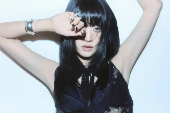 A promotional photo of MINNIE for her solo debut titled "Her". She is wearing a black sleeveless outfit that's sheer and reveals a black bra. The clothes matche her black hair. She's standing against a white background. Her arms are raised as she leans on the wall, one hand obstructing one of her eyes (she's also wearing blue lenses). She stares straight into the camera in a serious but also slightly sexy manner.