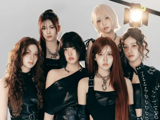 A photo of the 6 members of STAYC ("stay-see") on the set of the music video for BEBE. They're all dressed in black leather, with a lot of chrome accessories, buckles and jewelry. The background is white without much to see except for a big spotlight shining into the camera's direction. The girls are looking seriously/sexily into the camera, asserting a more adult identity than before.