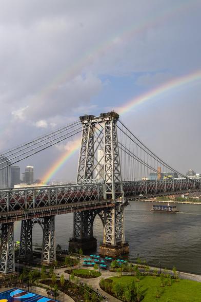 A view of a large suspension bridge and a park beside a large river. Behind the bridge can be seen half of a double rainbow, the outer arc much fainter than the inner one.