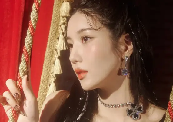A promotional photo for the album Lethality, showing Eun Bi standing next to a red curtain, holding the cord that will open or close it. Light is shining from what we can assume is the stage, into her eyes, as she looks into the distance. It's not clear what she's wearing, because it's taken from quite close, but her jewelry, including a four-leaf clover made out of dark blue gems, is very visible.