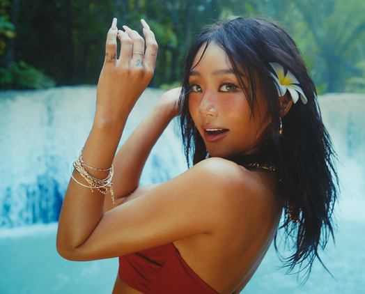 A promotional photo for Wait, showing Hyolyn with a deep tan (a rare thing in Kpop), her hair messy as if they had air dried, heavy eye makeup but only pink gloss on her lips, a large flower behind her ear, and wearing a dark red bikini. She's standing by a large tropical waterfall, some palm trees visible in the background above the water. She looks like she's dancing but also slightly surprised to, oh my God, I didn't see you there ! XD She looks at the camera over her shoulder, with her hands suspended in the air in a playful fashion, and a big side smile. It's the most attractive promotional photo I have ever seen in my life, if that helps you picture it.