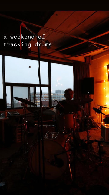 Photo of drummer behind drum kit, in silhouette in front of window at dusk with city buildings in background with smoggy sky