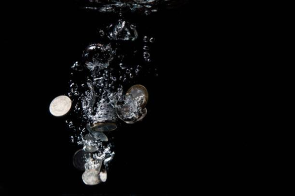 A cluster of silver coins plunges downward through dark water, trailing a flurry of air bubbles. The scene is captured mid-motion against a black background, emphasizing the reflective surfaces of the coins and the shimmering distortion of the bubbles. The overall effect is both dramatic and weightless, as the objects seem to float in a suspended moment of descent.