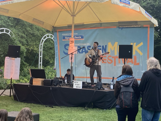 Ricky auf Bühne f. Ein junger Solo-Musiker auf einer Freilichtbühne im Park. Er spielt akustische Gitarre und singt dazu