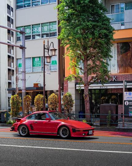 Ein roter Porsche 930 Turbo „Slant Nose“ steht bei Tageslicht an einer Ampel in Tokio, Japan. Das Fahrzeug ist mit klassischen Fuchs-Felgen ausgestattet. Im Hintergrund befinden sich moderne Gebäude mit großen Schaufenstern, ein Baum steht direkt am Straßenrand.