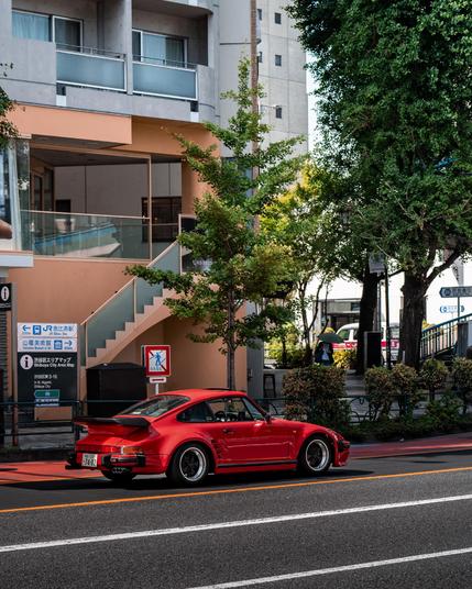 Ein roter Porsche 930 Turbo „Slant Nose“ steht bei Tageslicht an einer Ampel in Tokio, Japan. Das Fahrzeug ist mit klassischen Fuchs-Felgen ausgestattet. Im Hintergrund befinden sich moderne Gebäude mit großen Schaufenstern, ein Baum steht direkt am Straßenrand.