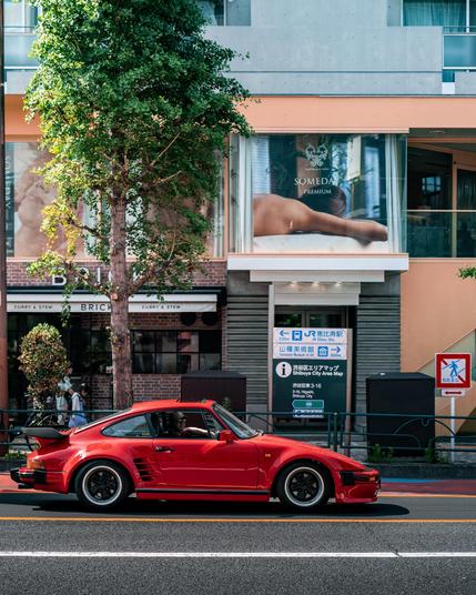 Ein roter Porsche 930 Turbo „Slant Nose“ steht bei Tageslicht an einer Ampel in Tokio, Japan. Das Fahrzeug ist mit klassischen Fuchs-Felgen ausgestattet. Im Hintergrund befinden sich moderne Gebäude mit großen Schaufenstern, ein Baum steht direkt am Straßenrand.