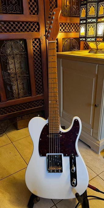 Une guitare électrique blanche, touche en érable, pickgard foncé, de face, dressée sur un stand.
C'est une copie de Fender Telecaster.