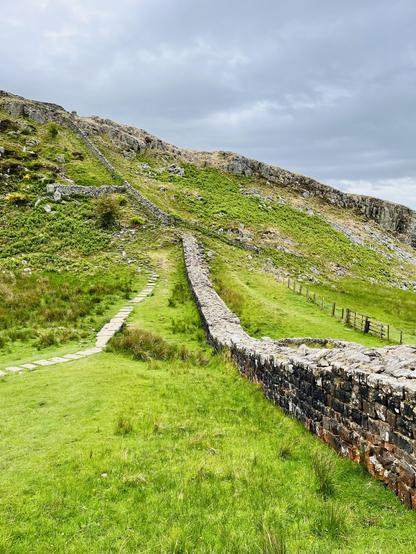 A view of Hadrian’s wall running along the right hand side of the image into the distance. A path runs nearly parallel on the left. The grass between is particularly green