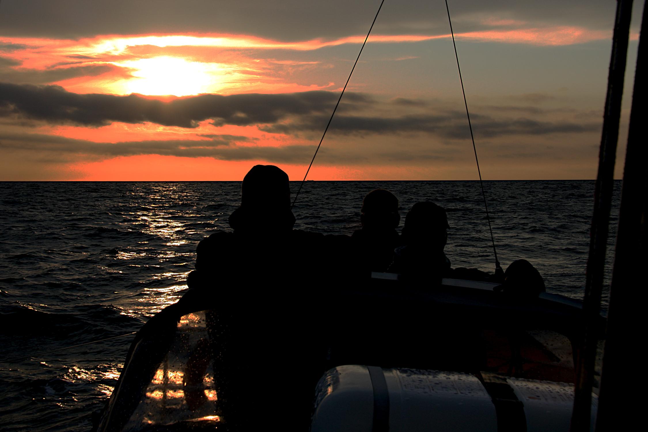 Drei Menschen stehen im Cockpit einer Segelyacht und schauen in die Kamera. Im Hintergrund ist die untergehene Sonne zwischen den Wolken zu sehen. Durch das Gegenlicht wirken die Personen wie Scherenschnitte. 
Die See ist ruhig, die Yacht hat leichte Krängung.