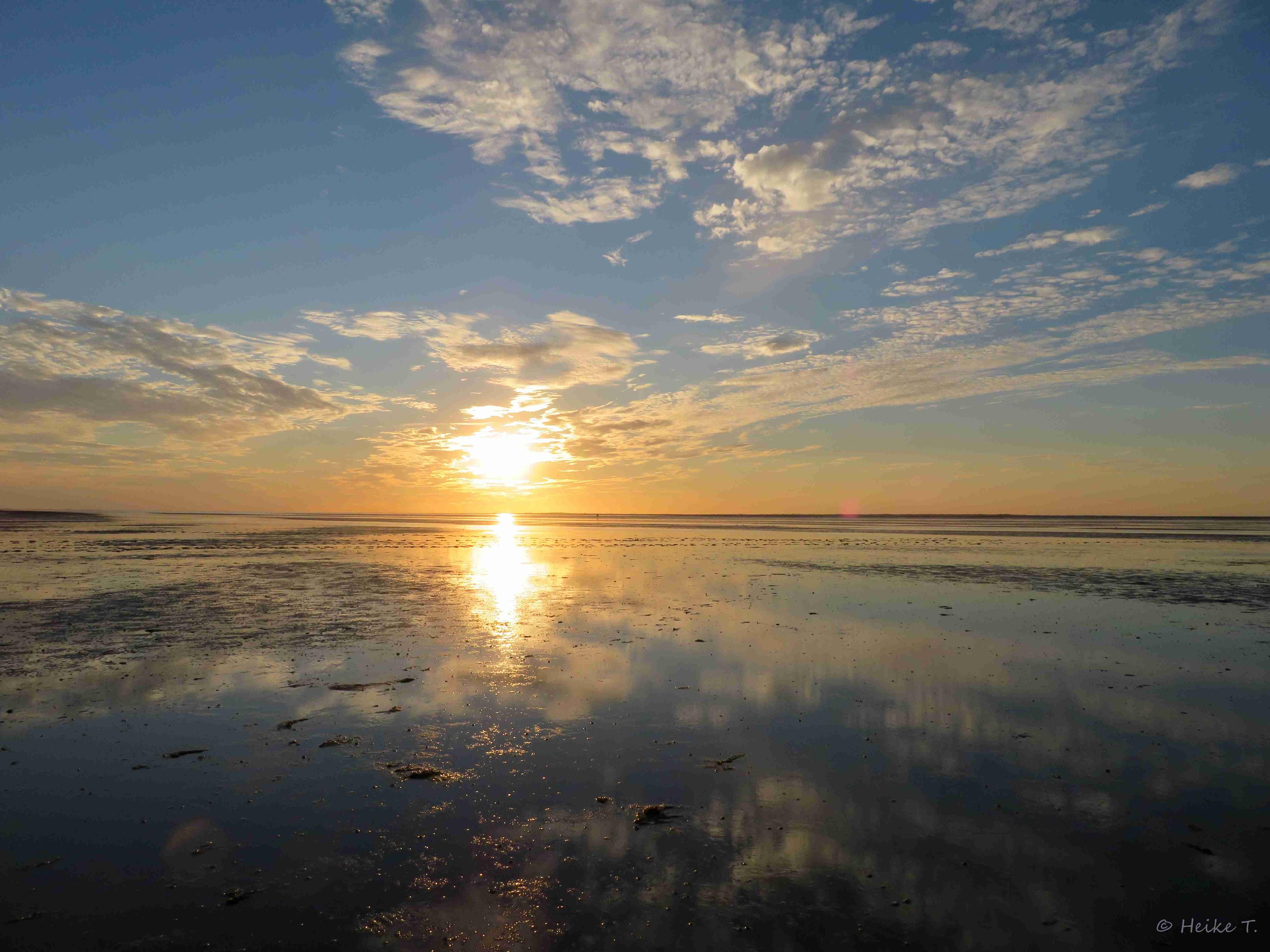 Die Sonne geht über dem Meer unter, es herrscht Ebbe. Verstreute Wolken am Himmel, die sich unten im Wasser spiegeln.