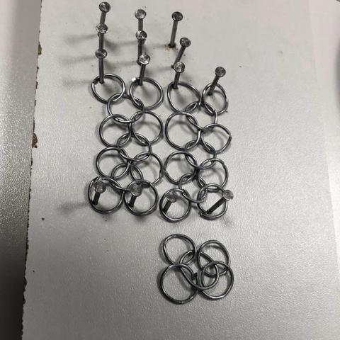 Several rows of chain mail being assembled on a jig made of nails in white laminated particle board. The rings a roughly circular, made of galvanized steel. A separate link of rice rings waits to be added. 