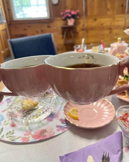 In the foreground two delicate teacups are tapped. In the background a muted  array of different goodies from an afternoon tea