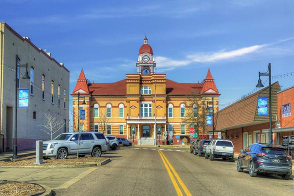 A historic courthouse with a red roof and clock tower stands prominently at the end of a street lined with parked cars.