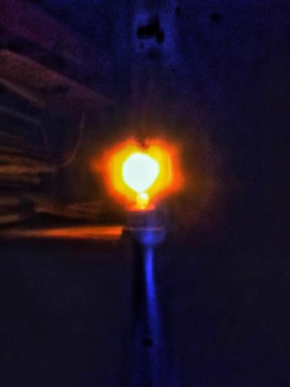La imagen muestra una vela encendida que emite una luz brillante y cálida de color naranja. La vela está montada en un soporte vertical, de madera, que se extiende hacia abajo. El fondo es oscuro, lo que hace que la luz de la vela sea aún más destacada. La imagen está ligeramente desenfocada, lo que sugiere que la foto fue tomada con una cámara que no estaba completamente enfocada o con una mano temblorosa. La luz de la bombilla crea un halo alrededor de su filamento, y se puede ver una pequeña cantidad de luz reflejada en la superficie del soporte. La atmósfera general es de un espacio oscuro y misterioso, con la vela como el único punto de luz.