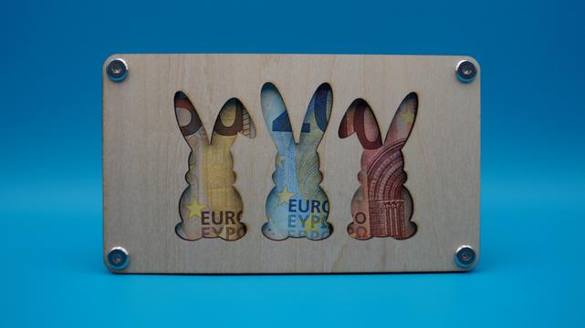 Verschlossener Holzrahmen mit drei Hasensilhouetten – dahinter leuchten bunte Euro-Banknoten hervor.