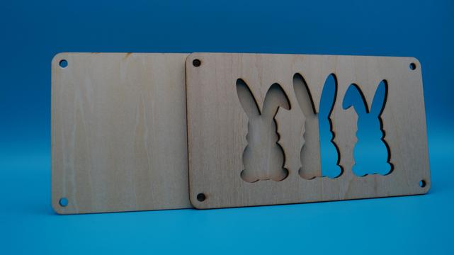Zweiteiliger Holzrahmen mit drei ausgeschnittenen Hasenformen, vorbereitet zum Einlegen eines Geldscheins.